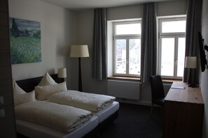 Une chambre d’hôtel avec deux lits, un bureau, une chaise, une lampe, un tableau et une fenêtre avec des rideaux.