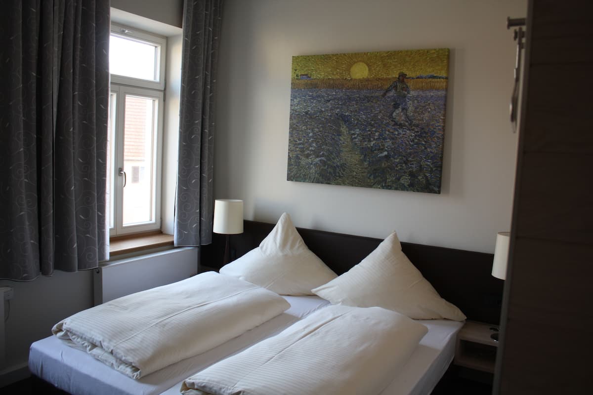 A hotel room with a bed, two bedside lamps, a window with curtains, and a painting on the wall.