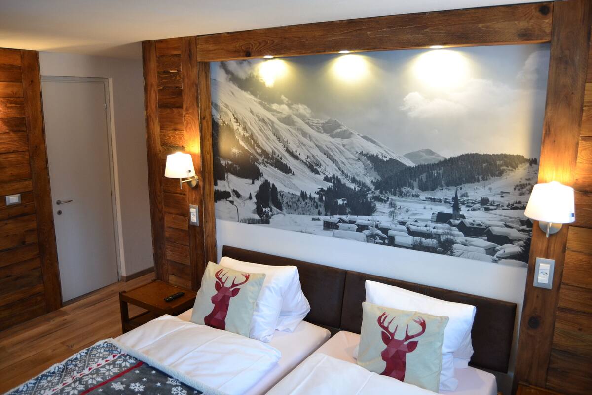 A hotel room with a large wall mural of a snowy mountain landscape, two beds with white bedding and deer-themed pillows, wooden paneling, and two wall-mounted lamps.