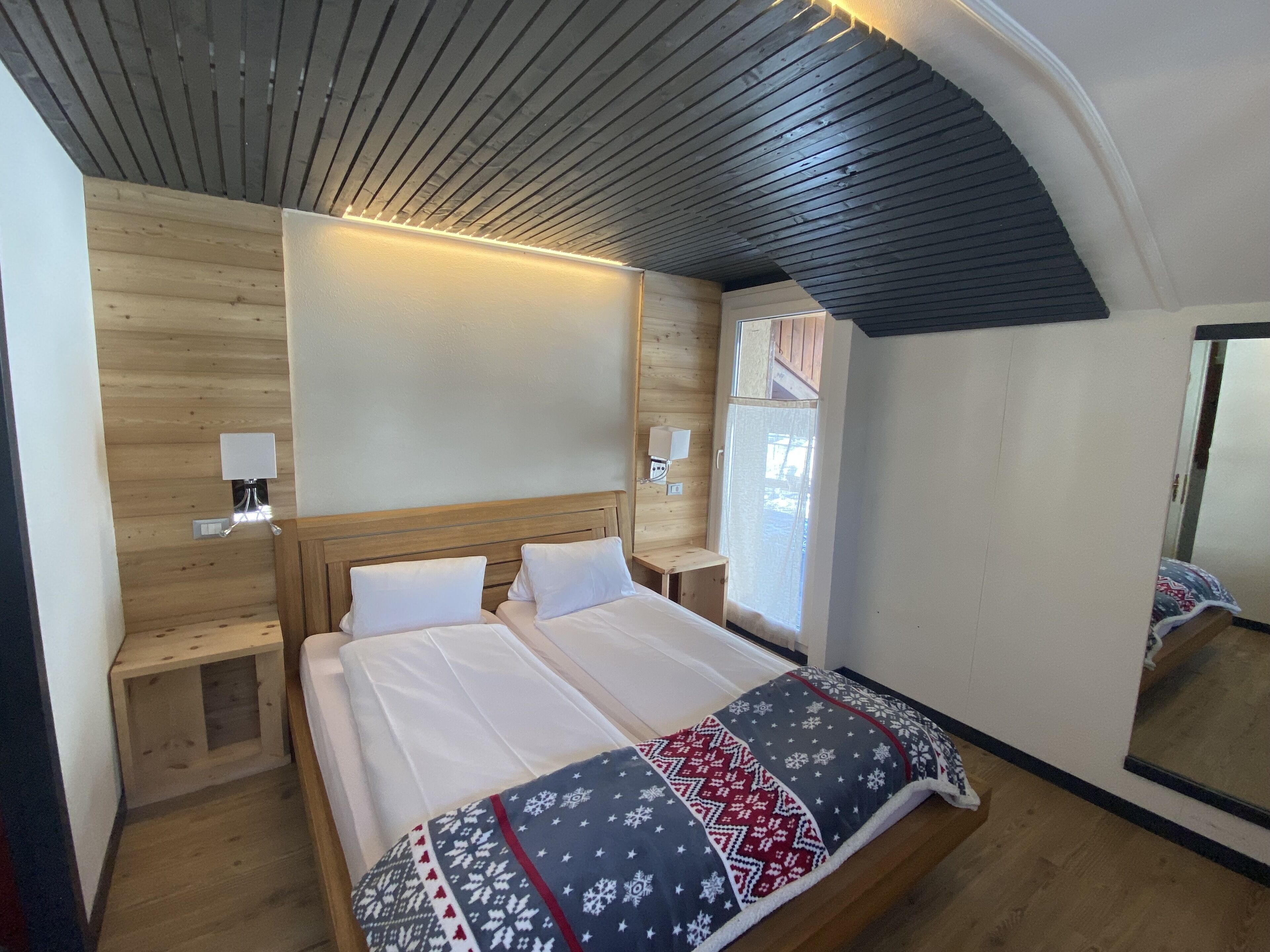 A bedroom with a wooden ceiling, a bed with white bedding, a patterned throw blanket, and a nightstand with a lamp.