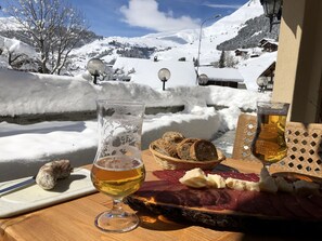 Uma mesa com um copo de cerveja, um prato de embutidos e pão, ao fundo uma paisagem montanhosa coberta de neve.