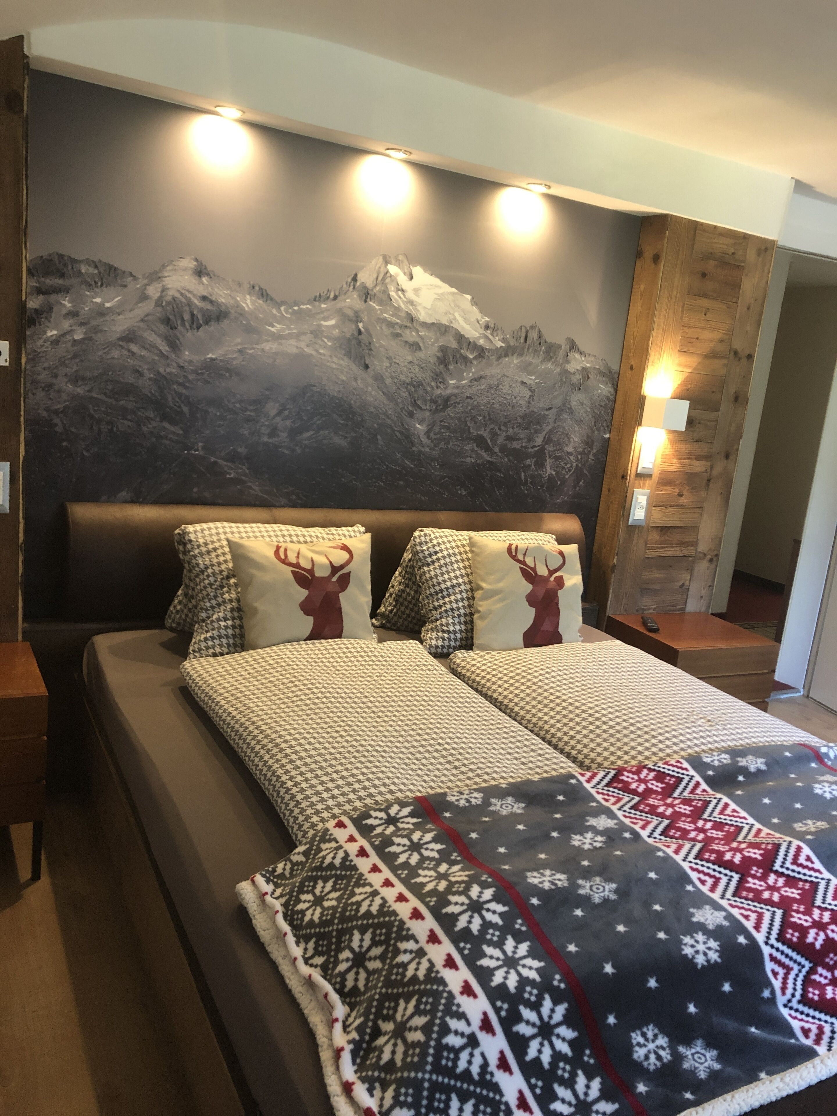A bed with a patterned bedspread and pillows, a mountain mural on the wall, and a wooden nightstand with a lamp.