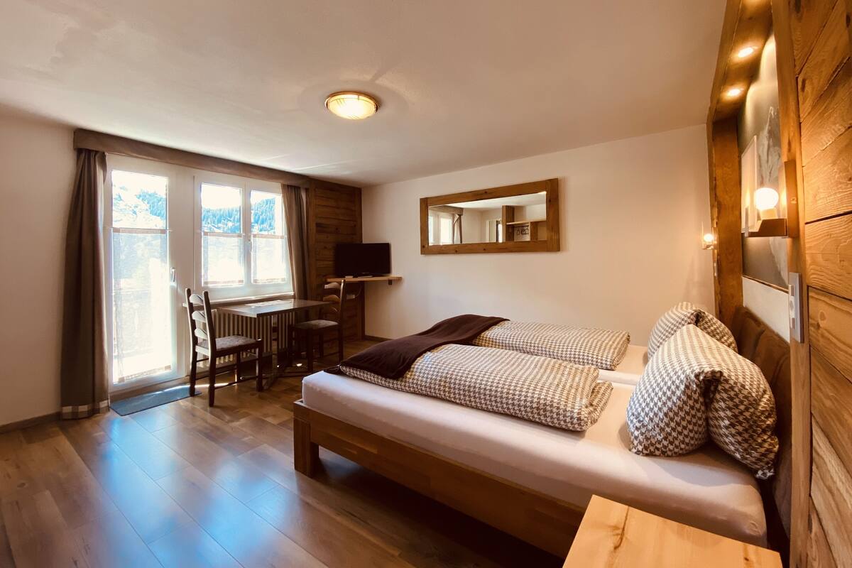A cozy room with a wooden floor, a bed with patterned pillows, a desk with a chair, and a television.