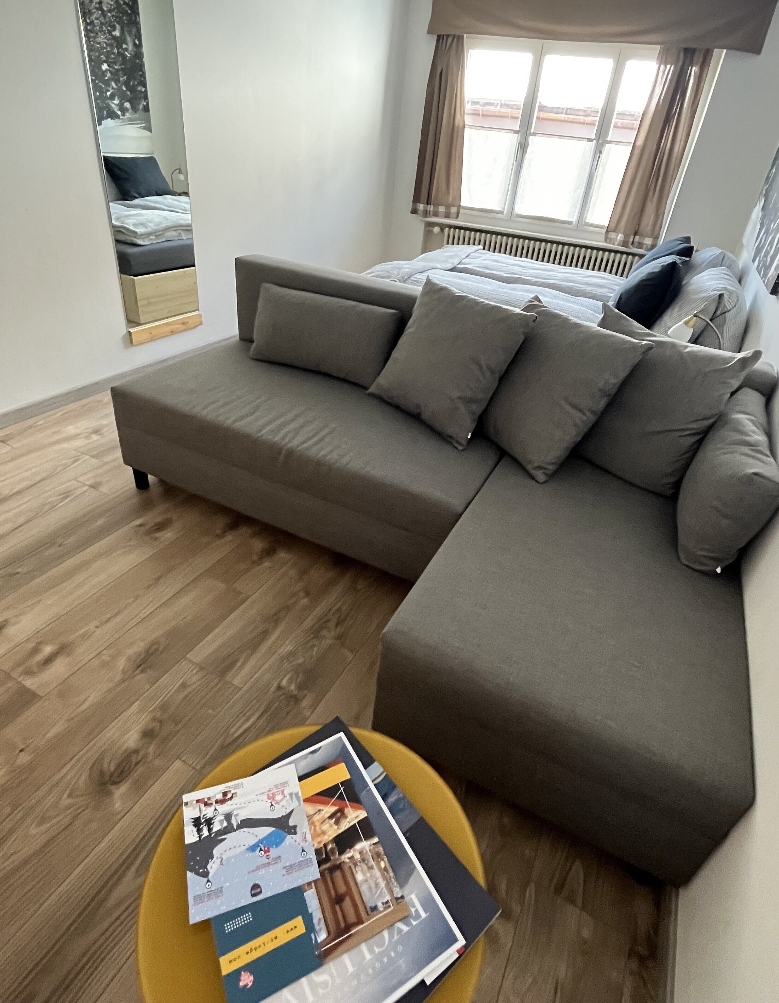 En moderne stue med en grå hjørnesofa, et rundt sofabord med magasiner og et vindue med gardiner.