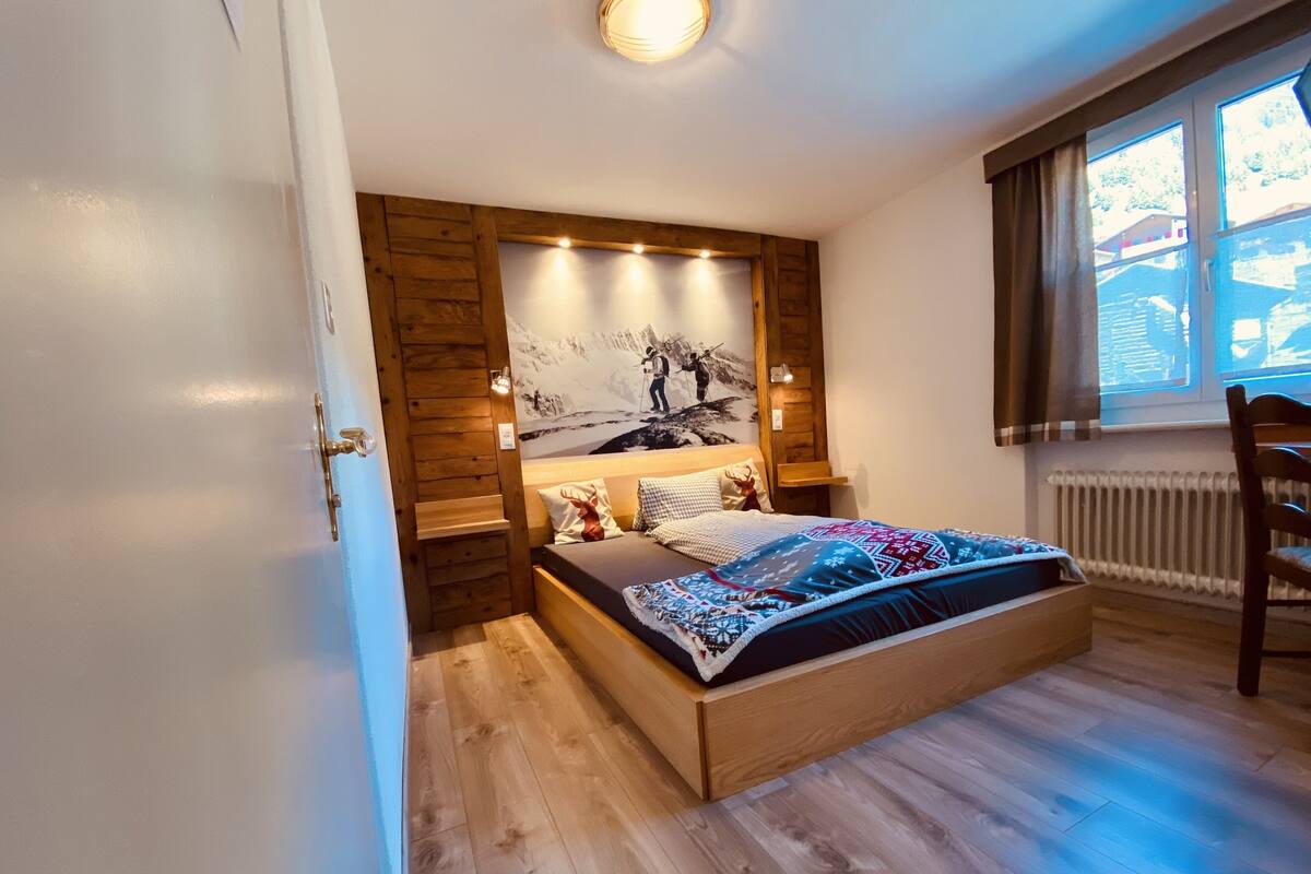 A bedroom with a wooden bed, a nightstand, a wall-mounted light, and a window with curtains.
