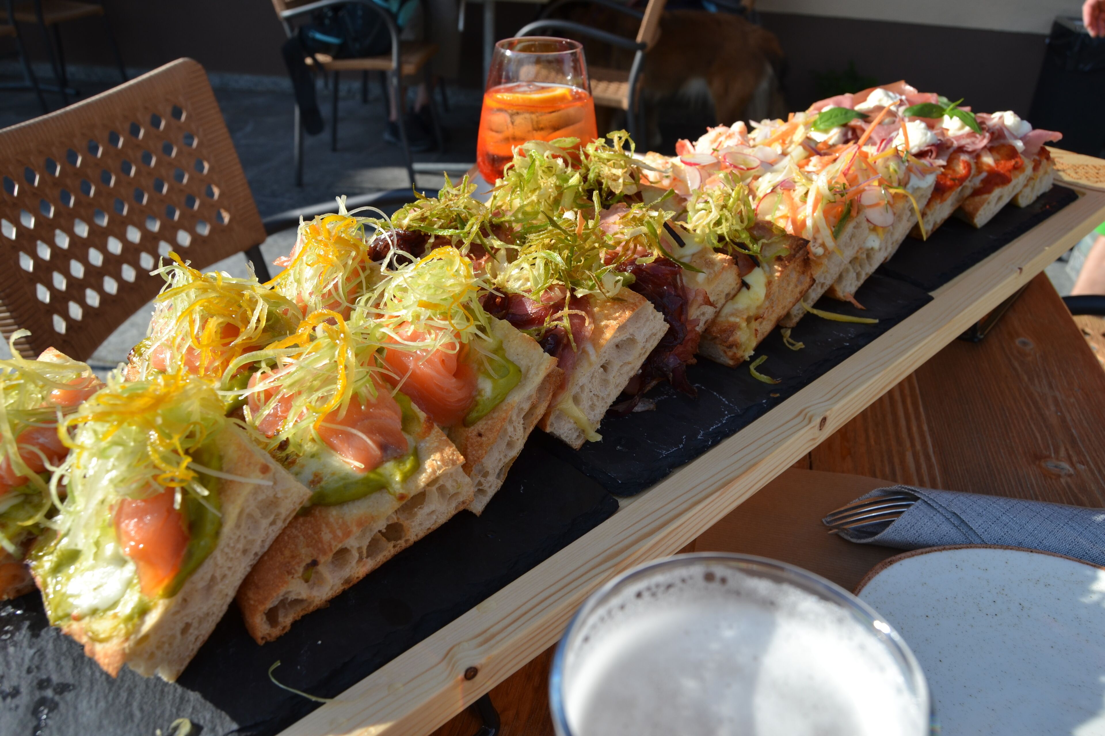 A wooden table with a black slate serving board holding a variety of open-faced sandwiches and a glass of iced tea.