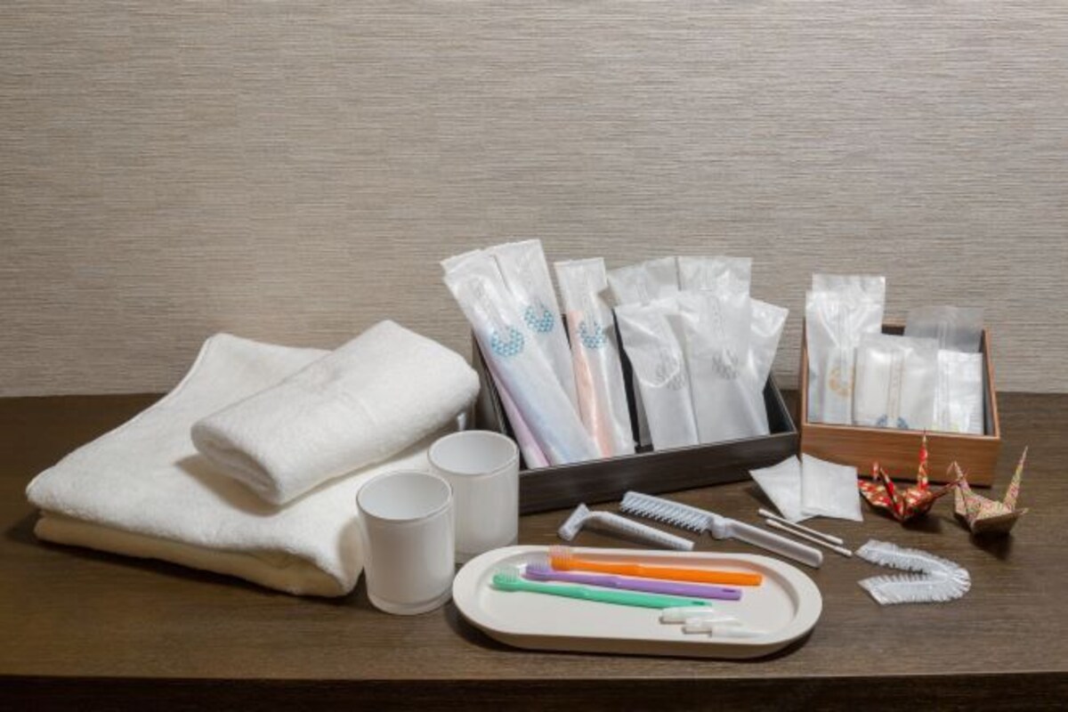 Ein ordentlich eingerichtetes Hotelzimmer mit einem Holztisch, auf dem Toilettenartikel, ein gefaltetes weißes Handtuch, zwei weiße Tassen und eine Ablage mit Zahnbürsten und einem Kamm ausliegen.