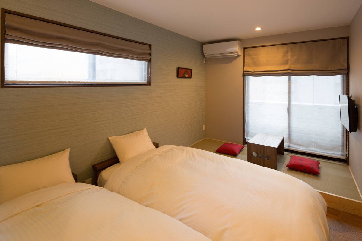 Een hotelkamer met twee bedden, een raam met gordijnen, een wandairconditioner en een klein tafeltje met een rood kussen.