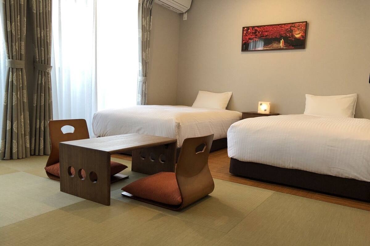 Een hotelkamer met twee bedden, een klein tafeltje en een stoel. Aan de muur hangt een schilderij en er is een raam met gordijnen.