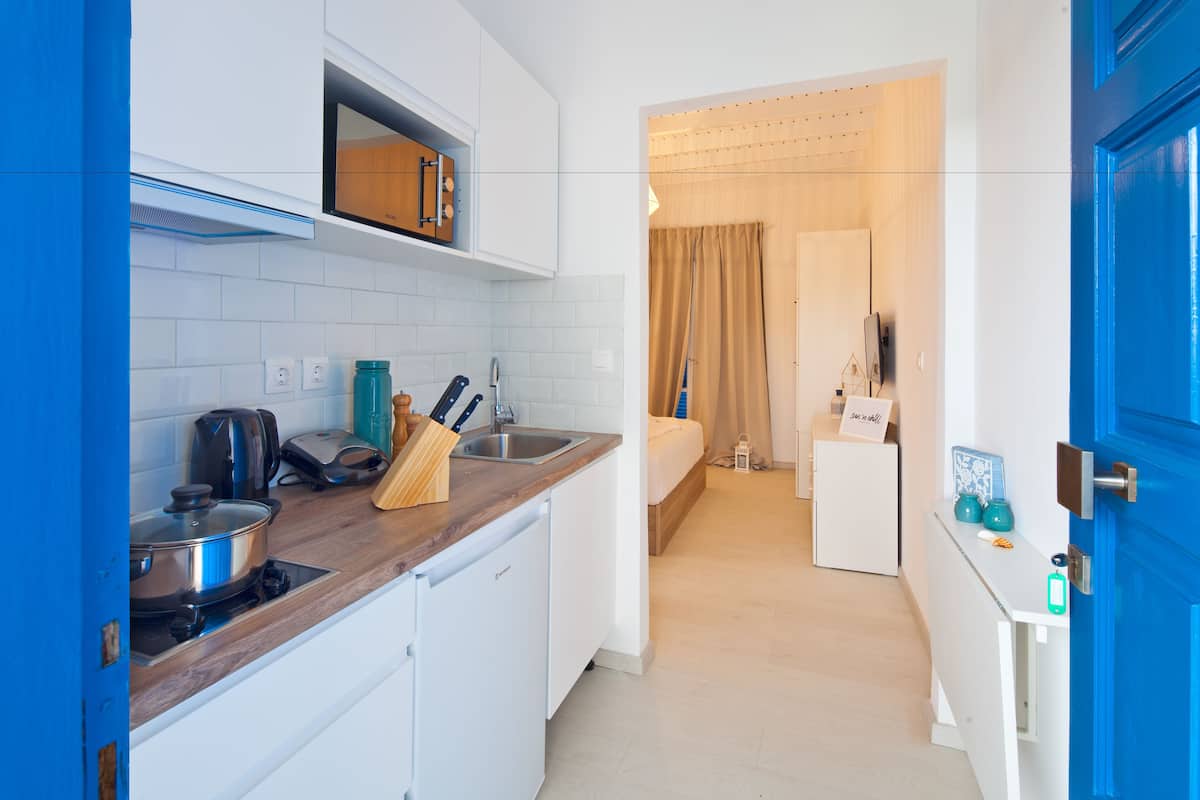 A compact kitchen with a stove, sink, and white cabinets. A bed is visible in the adjacent room, and a blue door is on the right.