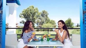 Two people sitting at a table with drinks and food, outdoors with trees and a clear sky in the background.