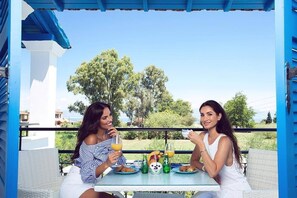 Two people sitting at a table with drinks and food, outdoors with trees and a clear sky in the background.