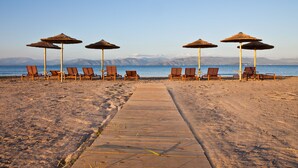 A beach with wooden lounge chairs and thatched umbrellas, a wooden walkway leading to the water, and mountains in the background. - Sun n Chill (Corfu)