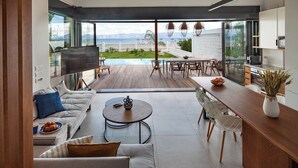 A modern living room with a large window, a pool, and a kitchen area. - Sun n Chill (Corfu)