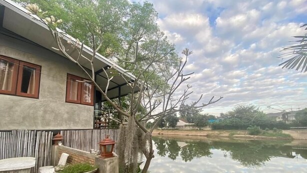 A house with a balcony overlooking a calm body of water, surrounded by greenery and a clear sky. - R-Lampang Guest House (Lampang)