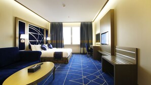 A hotel room with a bed, a sofa, a desk, a TV, and a large window with curtains. - Diwan Residence Hotel  Alsalamah (Jeddah)
