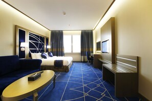 Habitación de hotel con cama, sofá, escritorio, televisión y un amplio ventanal con cortinas.
