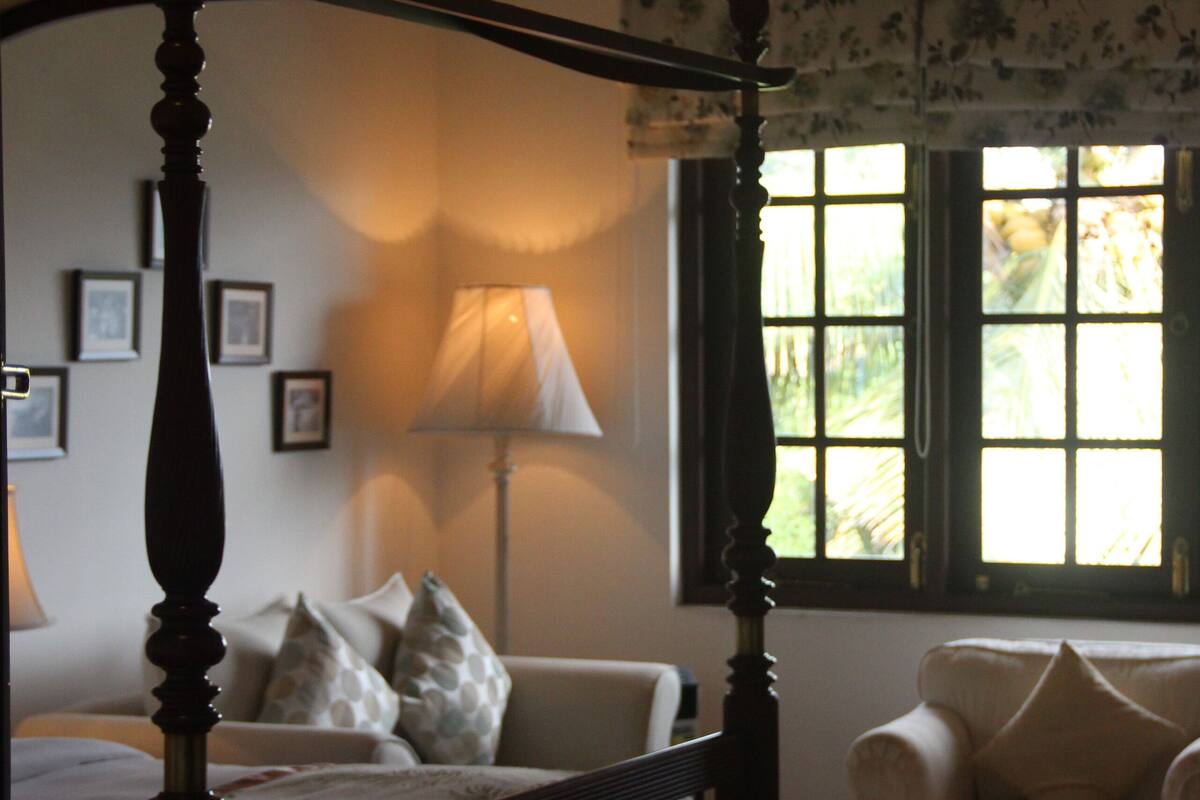 A bedroom with a four-poster bed, a sofa, a lamp, and a window with a view.