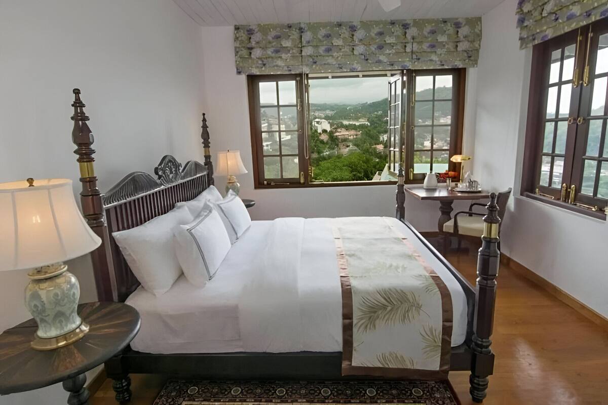 A neatly made bed with a view of a landscape through a window.