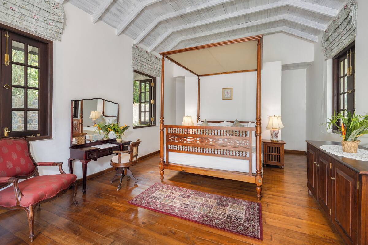A four-poster bed with a wooden frame, a desk with a mirror, a red upholstered chair, and a patterned rug.