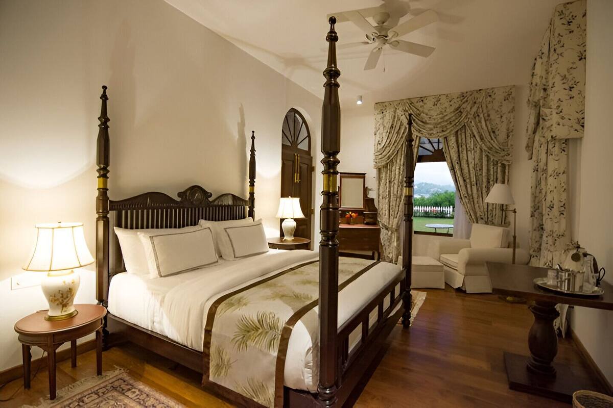 A four-poster bed with a wooden headboard, a bedside table with a lamp, and a view of the outdoors through a window with curtains.