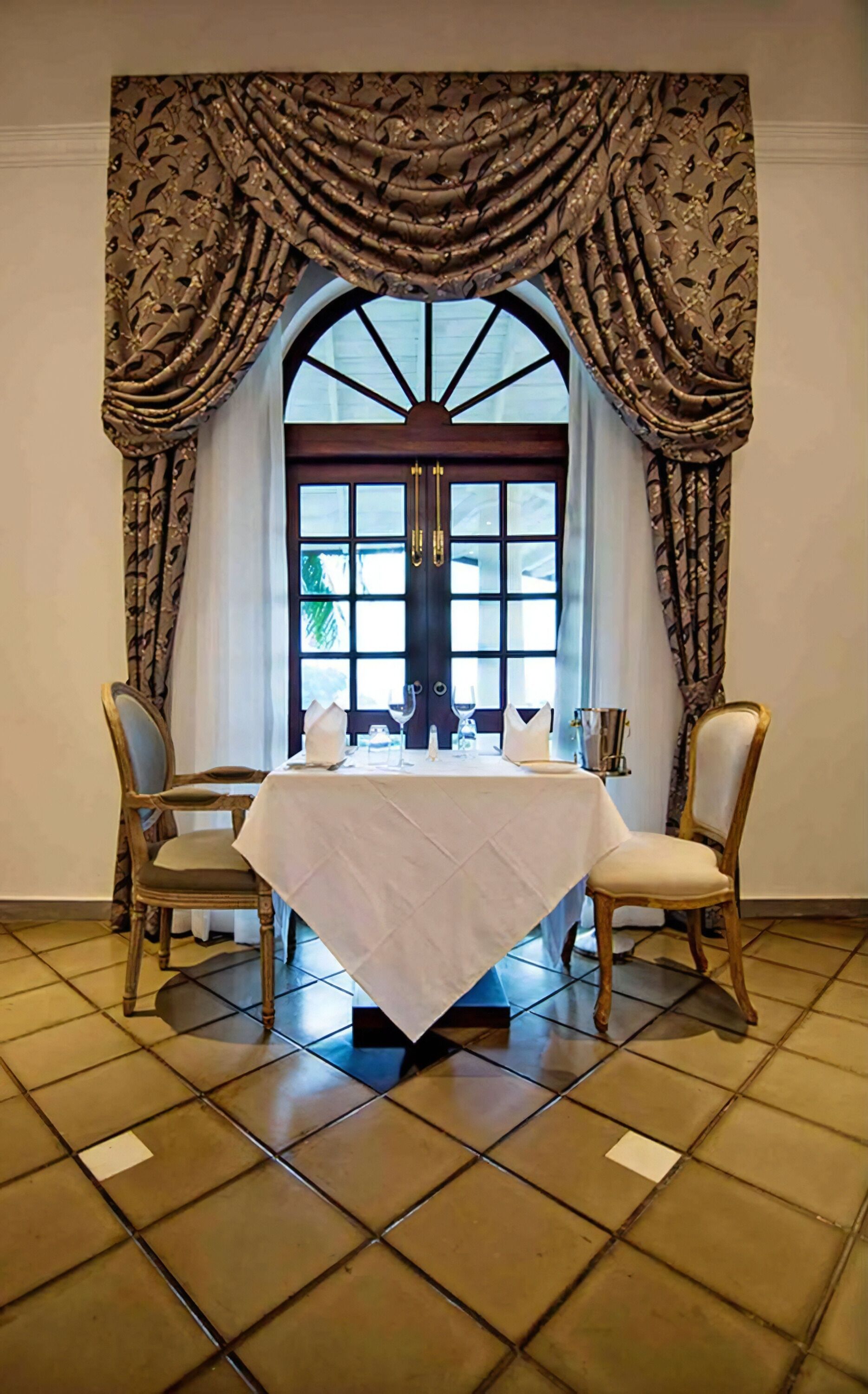 Un comedor con una mesa dispuesta para dos personas, sillas a cada lado y un ventanal con cortinas.