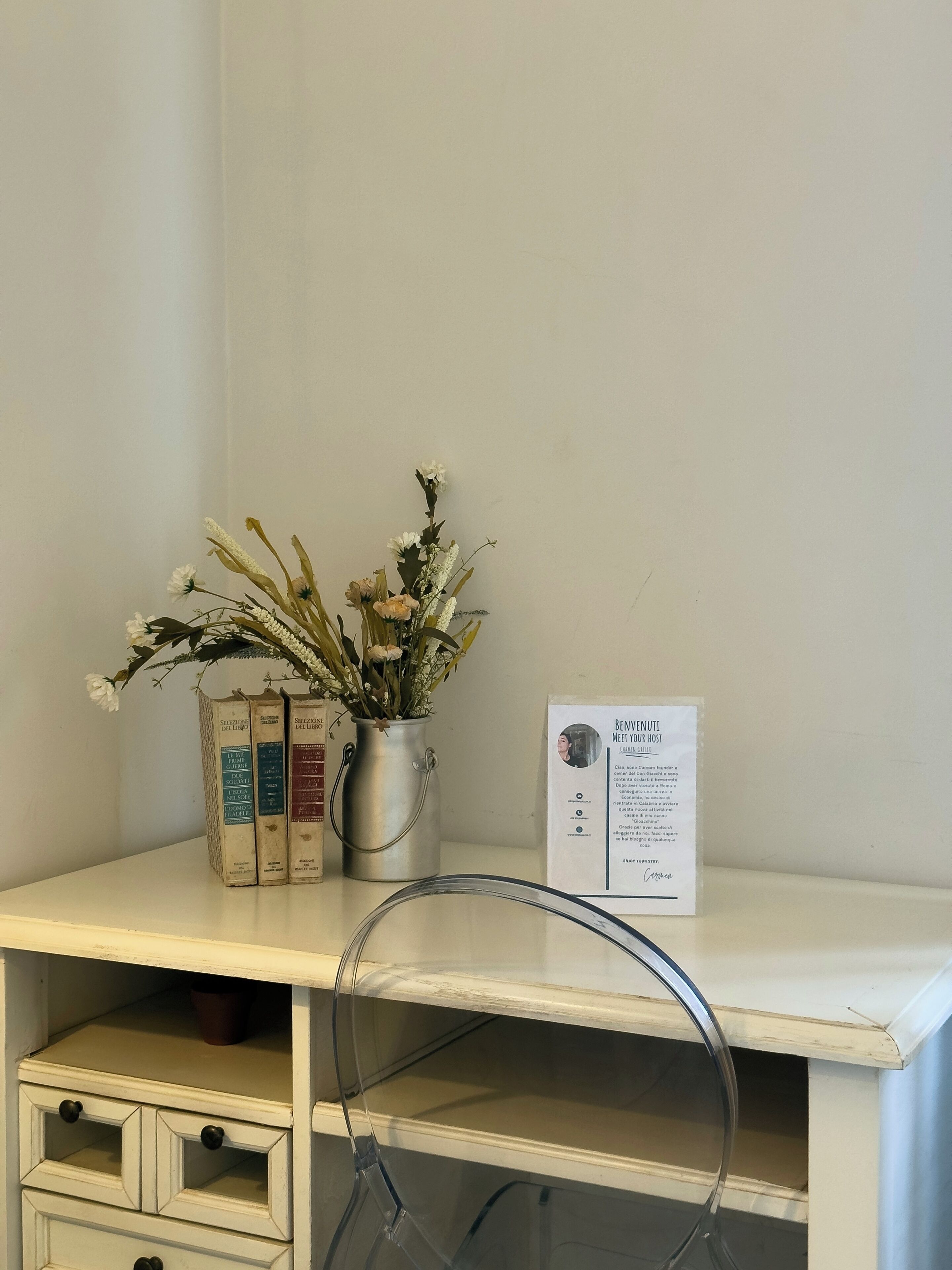 Un bureau blanc orné d’un vase en métal contenant des fleurs séchées, d’un récipient en métal rempli de livres et d’une affiche accrochée au mur.