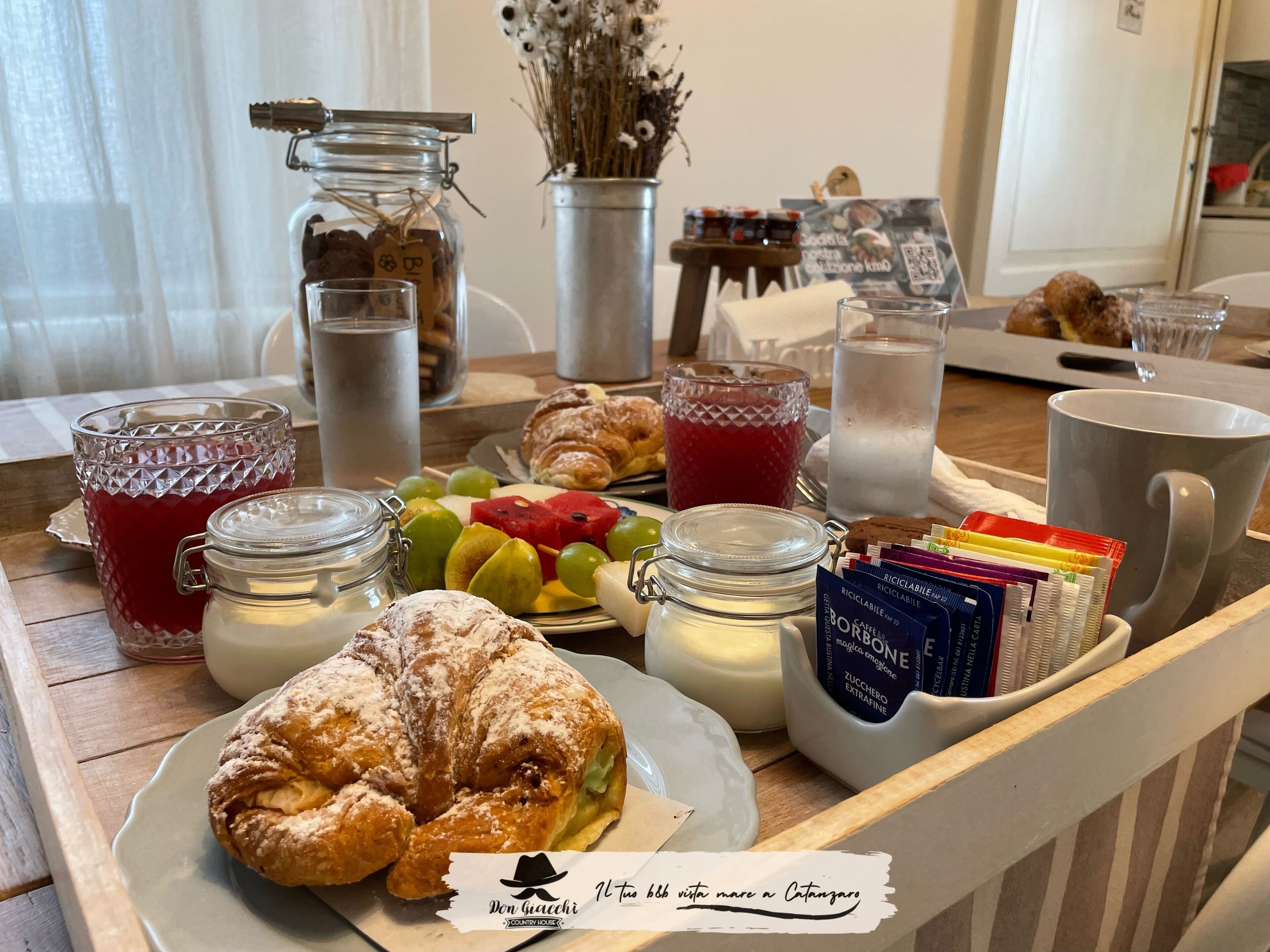 Una bandeja de desayuno con pasteles, frutas y bebidas, junto con una vela y un cartel con un código QR.