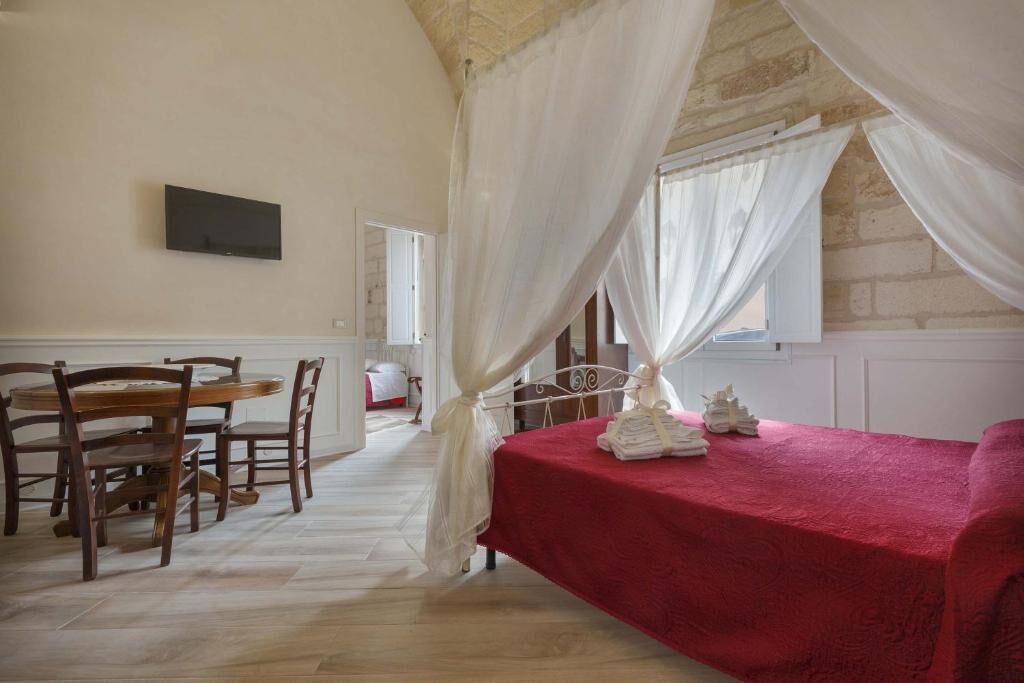 A room with a dining table and chairs, a bed with a red bedspread, and a white canopy over the bed.