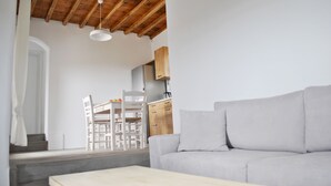 A modern living room with a light grey sofa, wooden ceiling beams, a dining area with a white table and chairs, and a kitchen in the background. - Livadi House Mykonos (Mykonos)