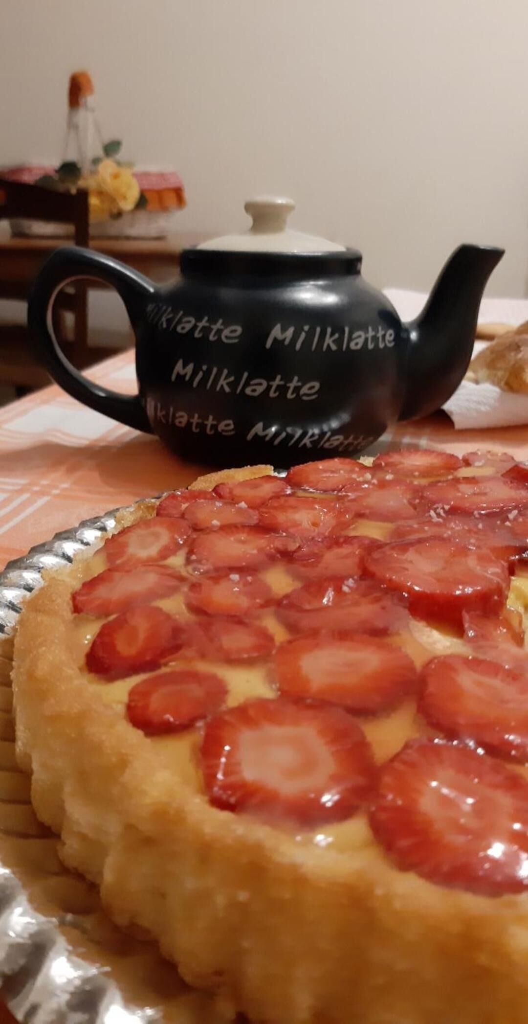 Eine schwarze Teekanne mit der Aufschrift "Milktatte Milchlatte", platziert auf einem Tisch mit einem Kuchen, belegt mit Erdrischeiben.