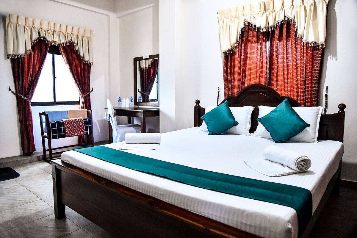 A hotel room with a large bed, two pillows, a wooden headboard, a desk with a chair, a mirror, and curtains.