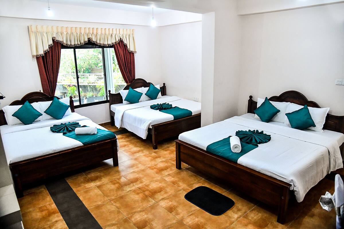 A hotel room with three wooden beds, each with white bedding and teal pillows. The room has a tiled floor and a window with curtains.
