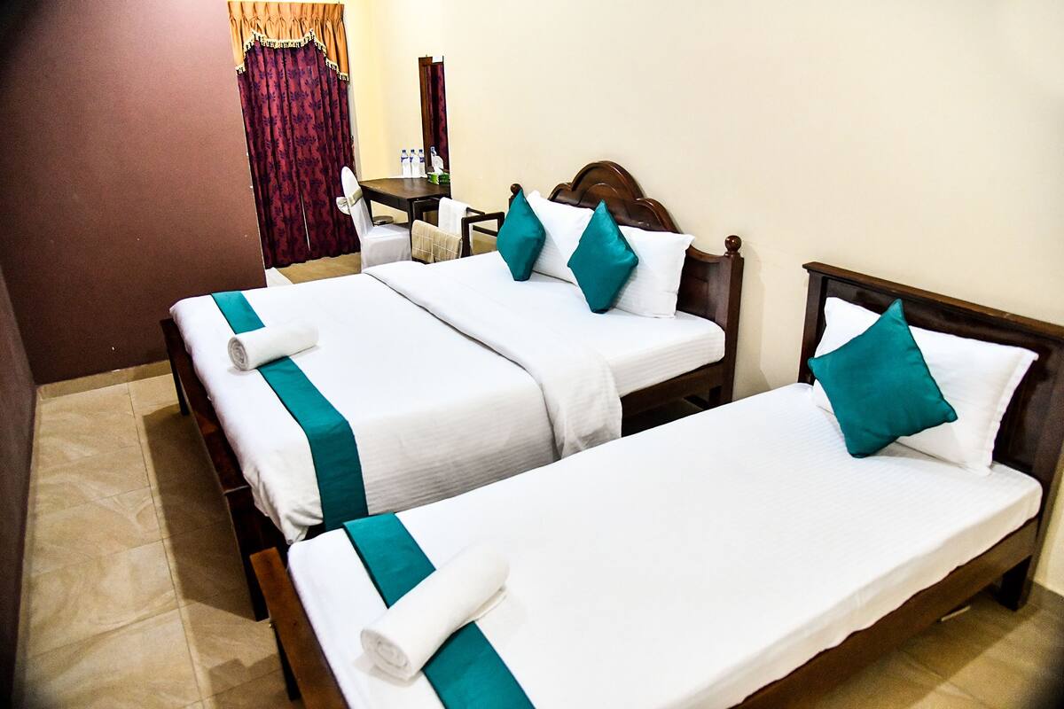 A hotel room with three single beds, each with white bedding and teal accents. There is a wooden headboard on the right bed, and a small table with a mirror and toiletries in the corner.