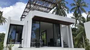 Ett modernt hus i två våningar med stora glasfönster, en träveranda och utsikt över palmer.
