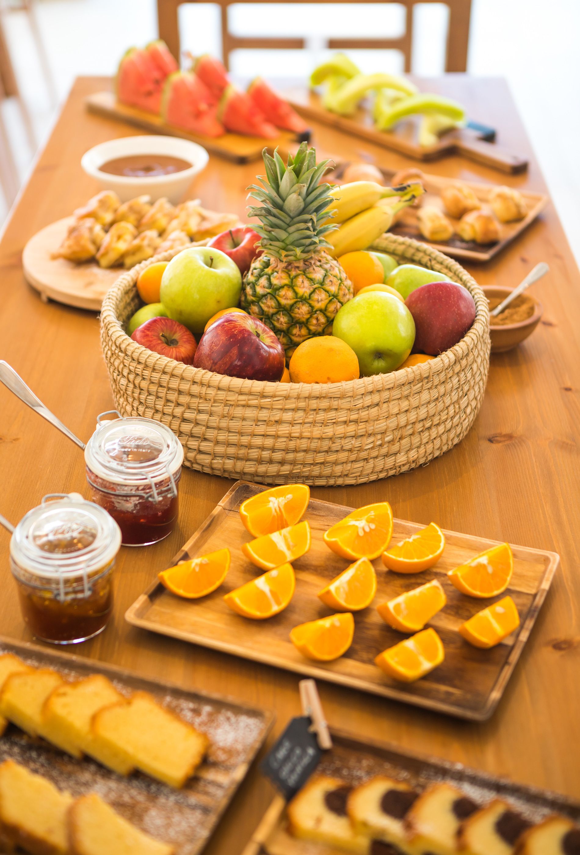 Una mesa de madera con una cesta de frutas que contiene manzanas, plátanos y una piña. También hay naranjas en rodajas y un frasco de mermelada.