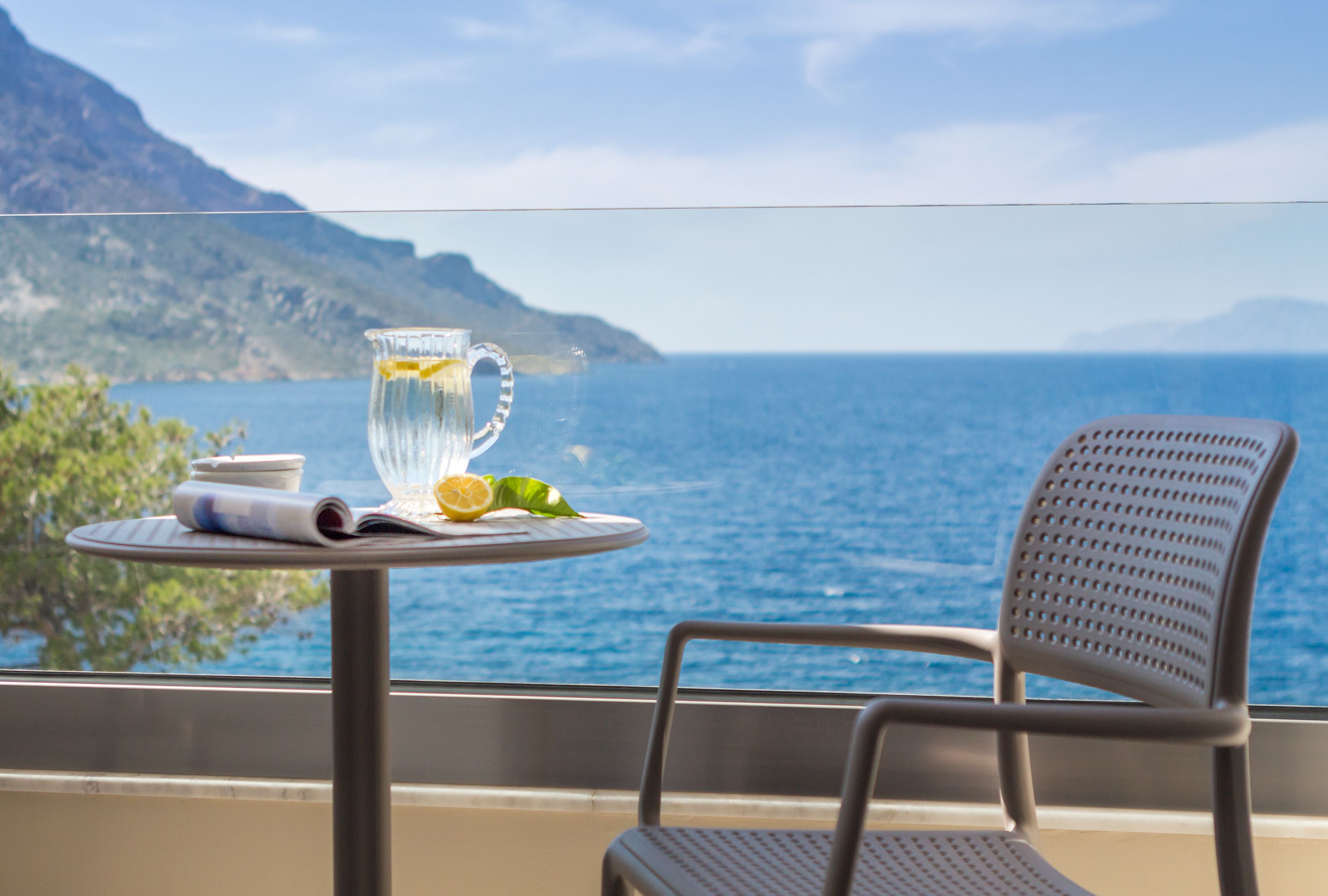 A table with a glass pitcher, a rolled-up menu, and a lemon on a balcony overlooking the sea.