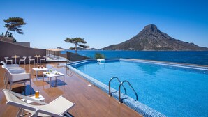 Zona junto a la piscina con sillones y mesas, vista despejada al mar y una montaña al fondo.
