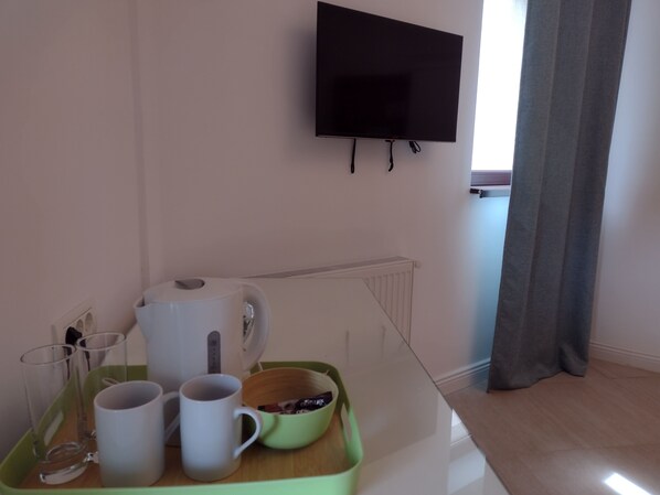 A room with a flat-screen TV mounted on the wall, a radiator, a wooden table with a tray holding glasses and mugs, and a window with curtains.