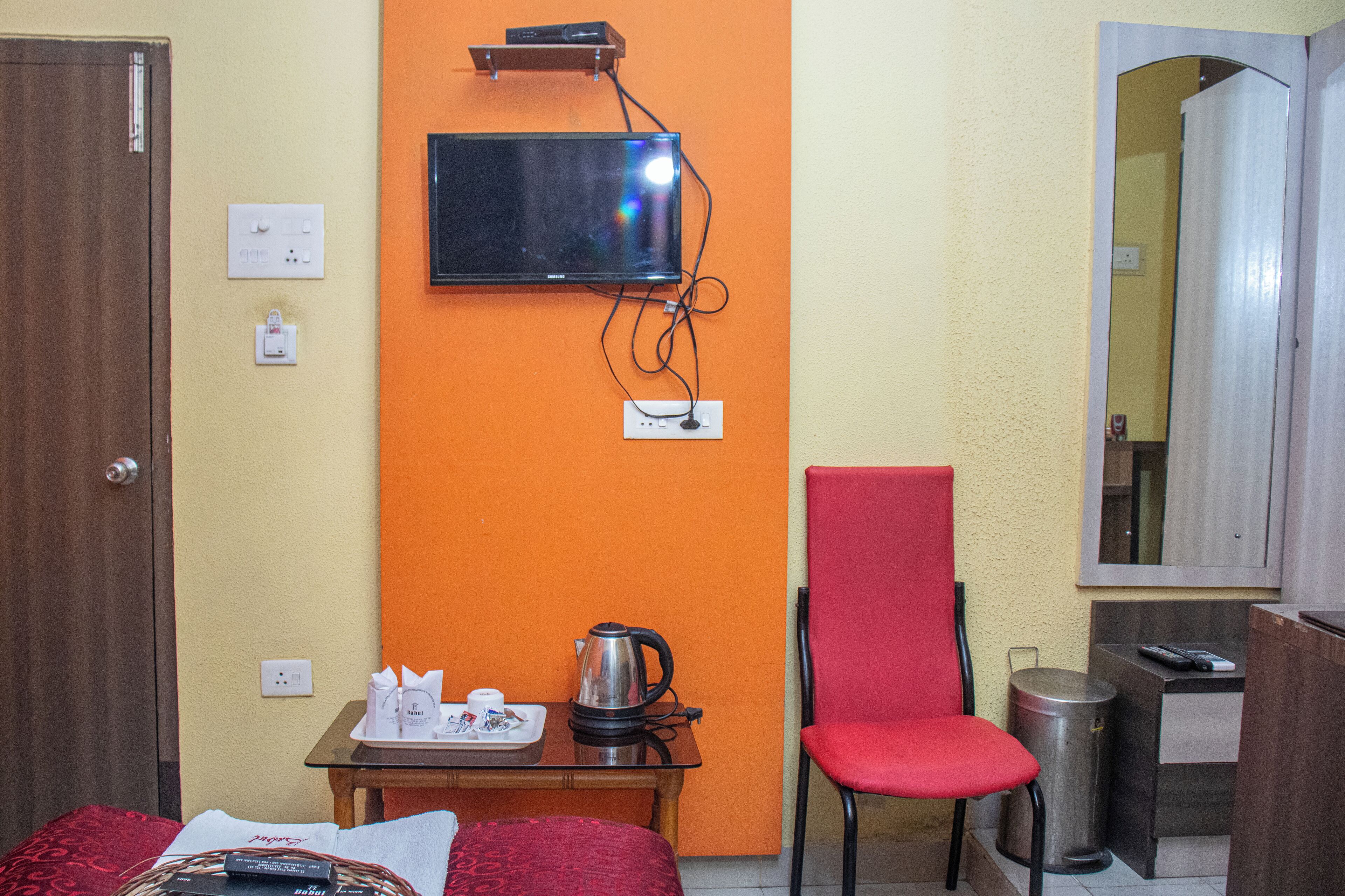 Habitación de hotel con cama, una mesa con hervidor, una silla, televisor montado en la pared y cubo de basura.