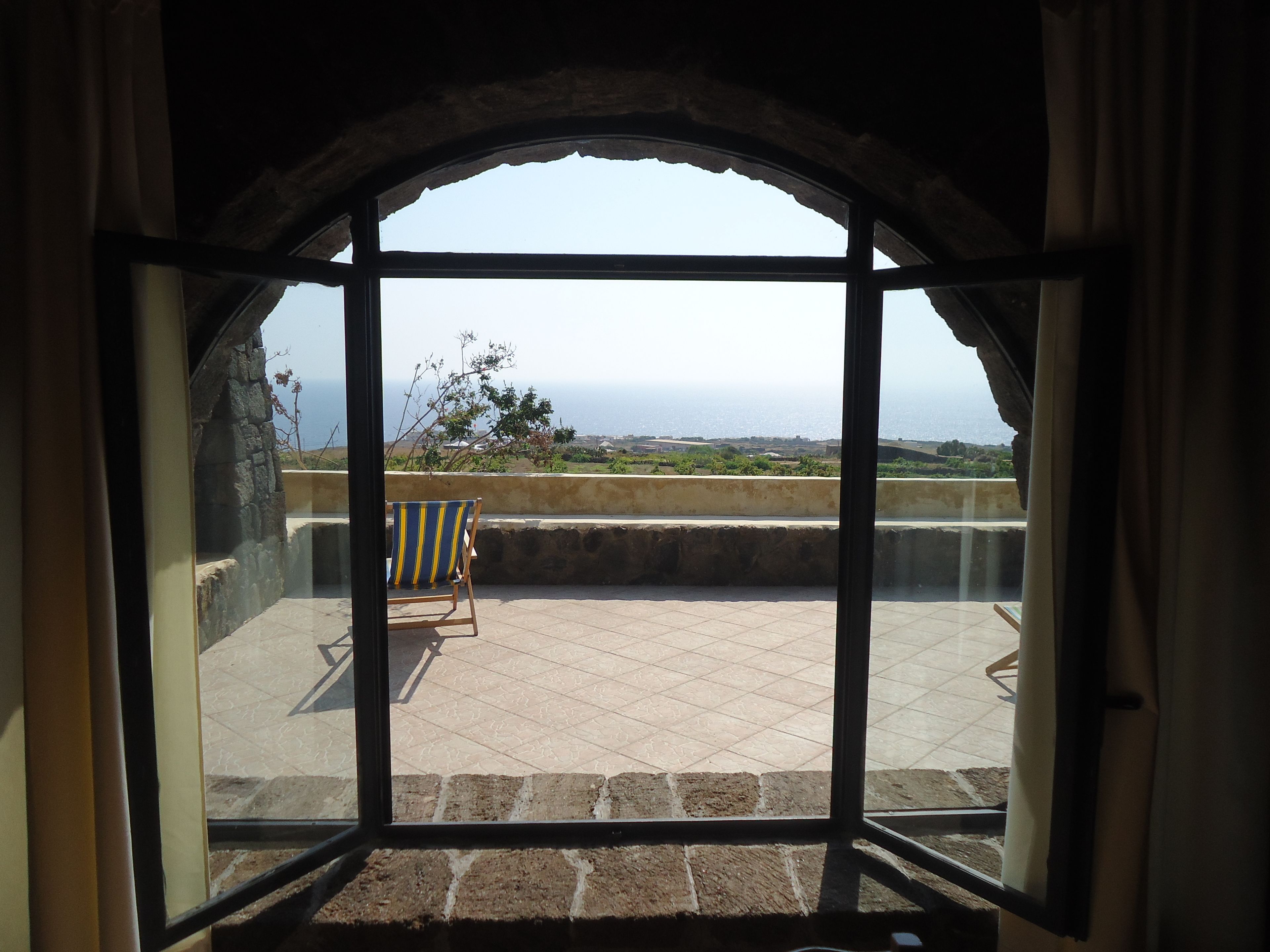 A view through a large arched window of a balcony with a chair, a stone wall, and a distant landscape.