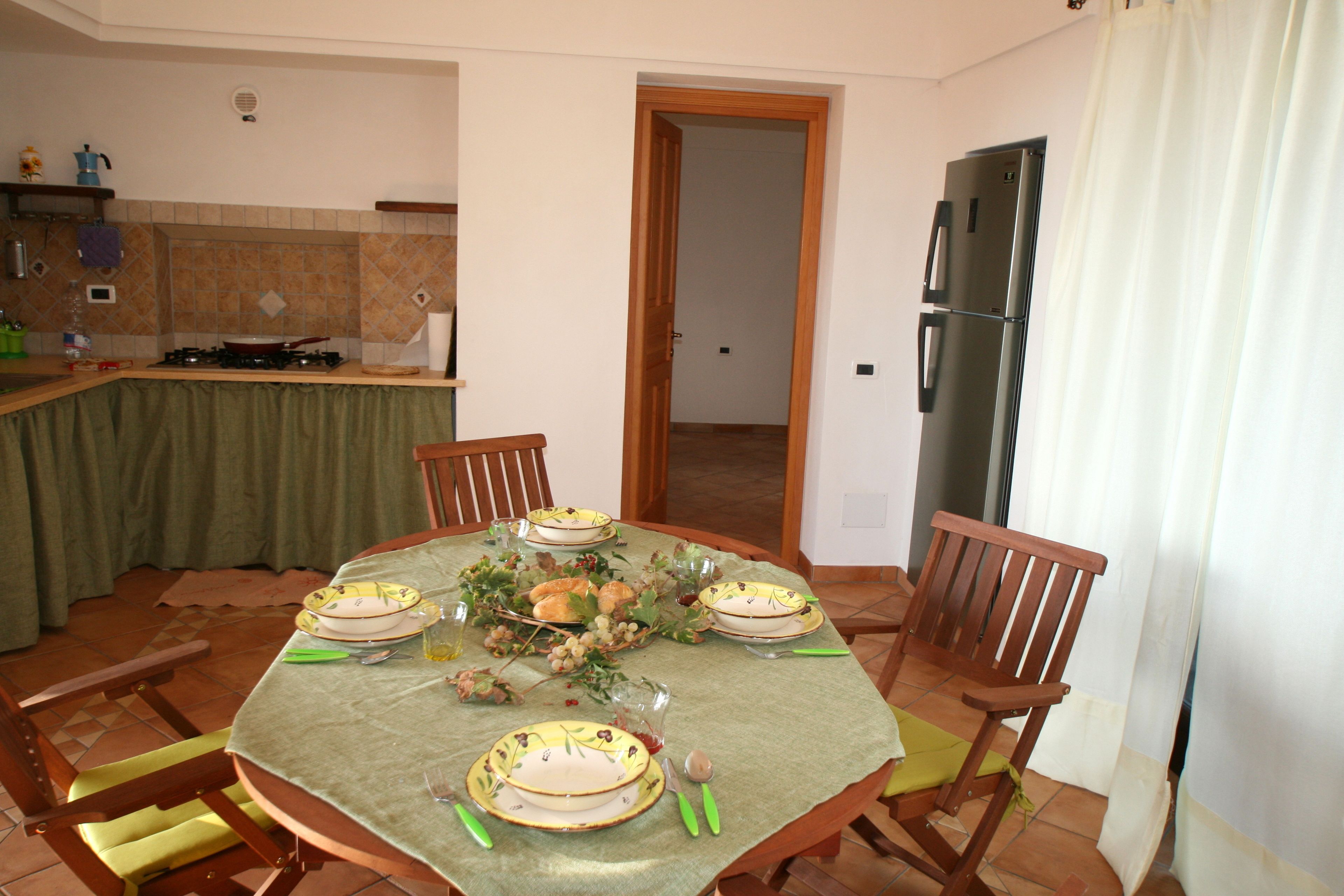 Un comedor con una mesa redonda preparada para cuatro personas, rodeada de sillas de madera. Se observa una cocina al fondo con un refrigerador y un lavabo.