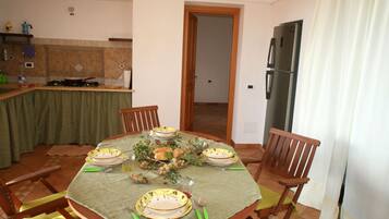 Un comedor con una mesa redonda preparada para cuatro personas, rodeada de sillas de madera. Se observa una cocina al fondo con un refrigerador y un lavabo.