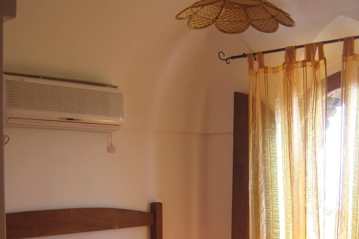 A bedroom with a wooden headboard, a striped bedspread, a small shelf with a glass, and a window with sheer curtains.