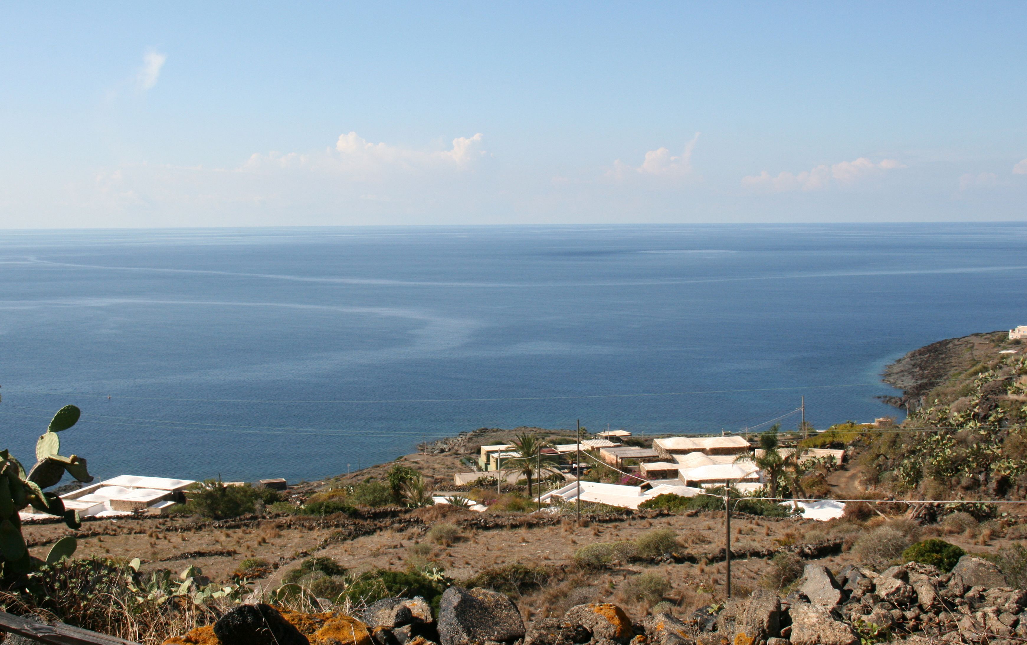 A coastal landscape with a view of the sea, rocky terrain, and a small settlement with buildings.