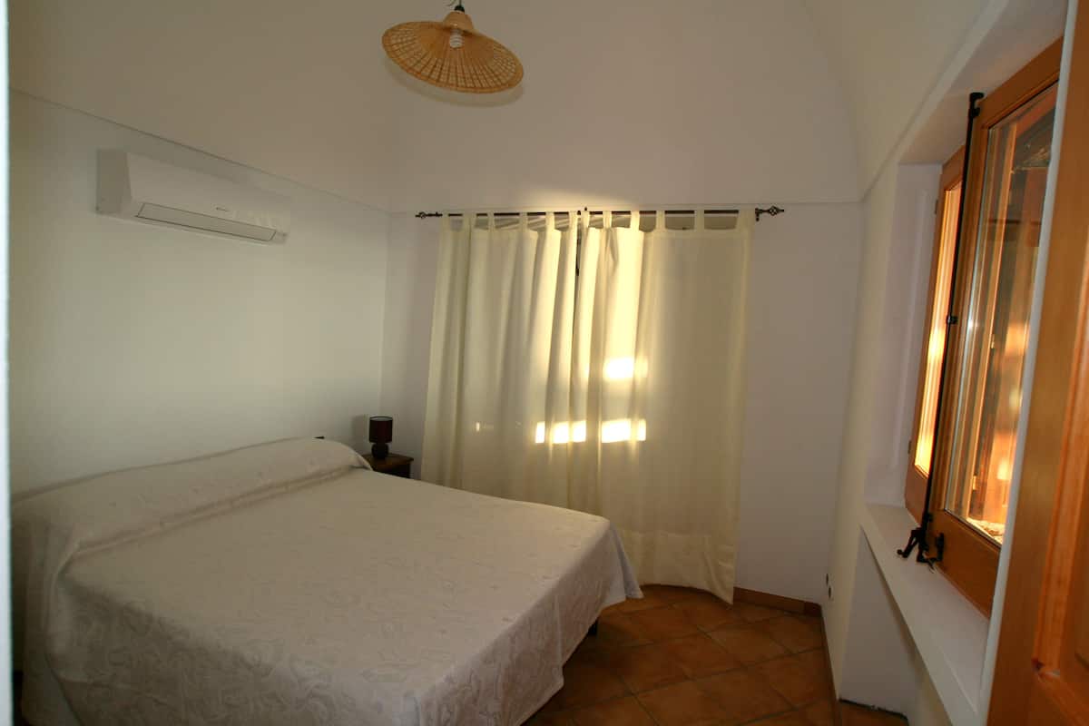 A bedroom with a bed, a window with curtains, a wall-mounted air conditioner, and a ceiling light fixture.