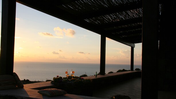 A sunset view from a covered outdoor area with a seating arrangement and a bottle on the table.