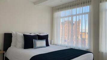 A hotel room with a large bed, white bedding, a dark blue throw blanket, and sheer curtains.