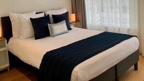 Una cama bien hecha con ropa de cama blanca y azul oscuro, una lámpara de noche y vistas de un paisaje urbano a través de cortinas traslúcidas.