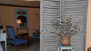 Una habitación con una puerta de madera rústica, una planta en maceta, un sillón tapizado azul, una mesita con lámpara y un espejo en la pared.